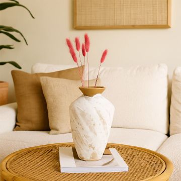 Handcrafted ceramic vase styled on a woven tray with dried pink florals in a cozy living room setting featuring neutral cushions warm textures and soft natural light.