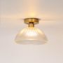 Ceiling light with brushed gold base and ribbed clear glass dome shade glowing on plain white background.