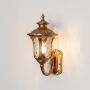 Gold metal outdoor wall light turned on against white background highlighting warm glow clear glass panels and decorative traditional lantern design.