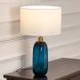 White shade teal glass table lamp on wooden sideboard with light on and white panel wall background for modern stylish décor.