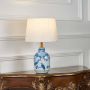 Blue and white ceramic table lamp with bird and floral detailing and a cream fabric shade illuminated against a white background.