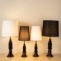 Four table lamps with carved wooden bases and fabric shades in white and black placed on a light surface against a textured light wall.