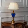 Blue glass table lamp turned on with pleated fabric shade and brass detailing displayed on white background.
