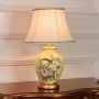 Yellow ceramic table lamp featuring floral bird hand painting gold metal base and fabric shade displayed on white background.