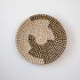 Handwoven wicker basket wall decor in beige with black and white swirl woven pattern on clean white background.