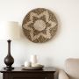 A round woven wall basket with black and beige spiral detail hangs above a dark table with a lamp and white ceramics on a tray near a chair.