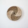 Handwoven wicker basket wall decor in beige with black spiral woven pattern design on clean white background surface.