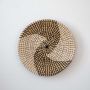 Handwoven wicker basket wall decor in beige with black and white spiral woven pattern design on clean white background.
