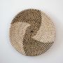 Handwoven wicker basket wall decor in beige with black and white spiral woven geometric pattern on clean white background.