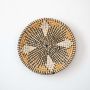 Handwoven wicker wall plate in black and natural tones on plain white background creating eco friendly decorative basket wall art.