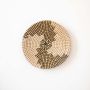 Handwoven wall basket in natural and black fiber with abstract swirl design shown on white background.