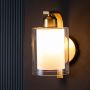 Gold wall sconce with frosted glass shade illuminated against a dark background showing warm light reflections and emphasizing the modern curved arm silhouette.