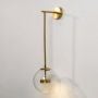 Gold wall light with clear glass globe shown switched on against a white background casting a gentle warm glow through the transparent round shade.