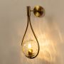Brushed gold teardrop wall light glowing on white background showcasing warm illumination and artistic curved frame with hammered glass globe.