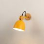 A yellow Scandinavian wall light with a matte dome shade and wooden mount shown with the light on against a white background giving a warm modern glow.