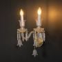 Double arm crystal wall light with gold base ornate glass design and crystal drops glowing on black background.
