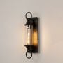 Black metal wall sconce with clear glass tube illuminated warmly on white background for inviting outdoor lighting.